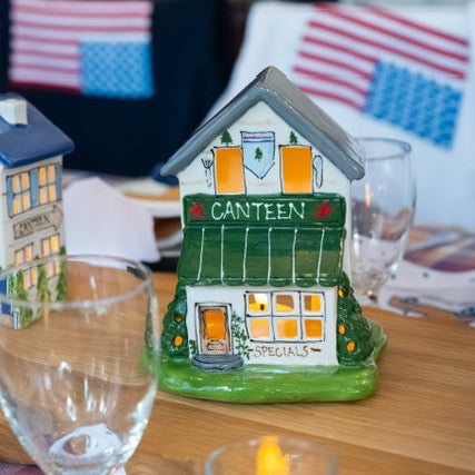 Decorative house-shaped village house with 'Canteen' on a table with glasses and a candle.